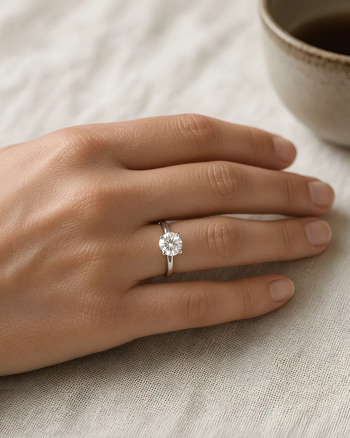 Round brilliant lab-grown diamond solitaire in a four-prong white gold setting on the left ring finger, resting on ivory linen